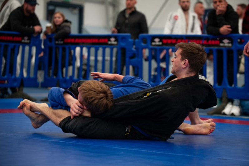 Two people in a BJJ match, one in a black gi and one in blue. The person in black seems to be performing a Bow-And-Arrow choke on the other person